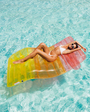 Woman relaxing on a colorful FUNBOY lounger pool float in clear blue water, wearing sunglasses and a white swimsuit.